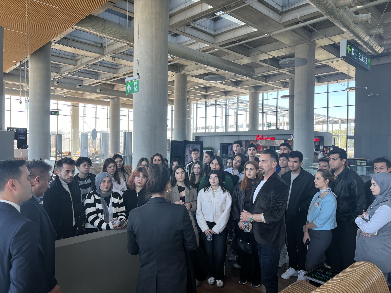 This photo shows the group gathered inside a terminal, listening to a representative in a navy blue suit. The interior has a striking architectural design with raw concrete columns and a ceiling equipped with square skylights. There are modern, wave-shaped wooden benches on the right.