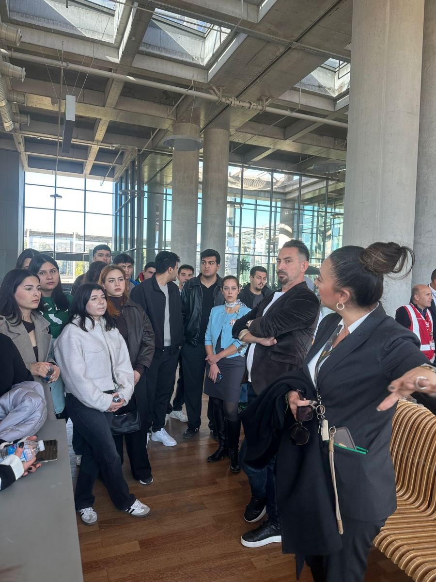Another scene from inside the terminal shows the female official in a black suit continuing her presentation among large concrete pillars. Airport signage for gates 31A-31B and 32 is visible, and the floor is made of polished wood. Bright daylight streams in through the large glass windows in the background.
