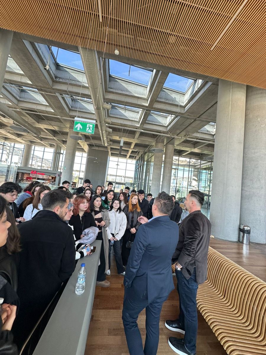 Taken from a different angle within the terminal, this image shows a female official in a black suit gesturing while explaining something to the group. A staff member from Çelebi, wearing a turquoise uniform and scarf, is also present among the students. The group appears focused and engaged in the technical visit. 