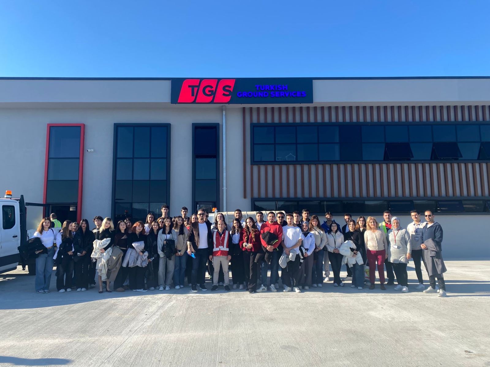 The group of students is posing in front of a modern, grey building with a large "TGS Turkish Ground Services" sign. The building features large black-framed windows and a section with vertical wooden slats on the facade. A staff member in a red safety vest is standing in the middle of the group, and the weather is bright and sunny.