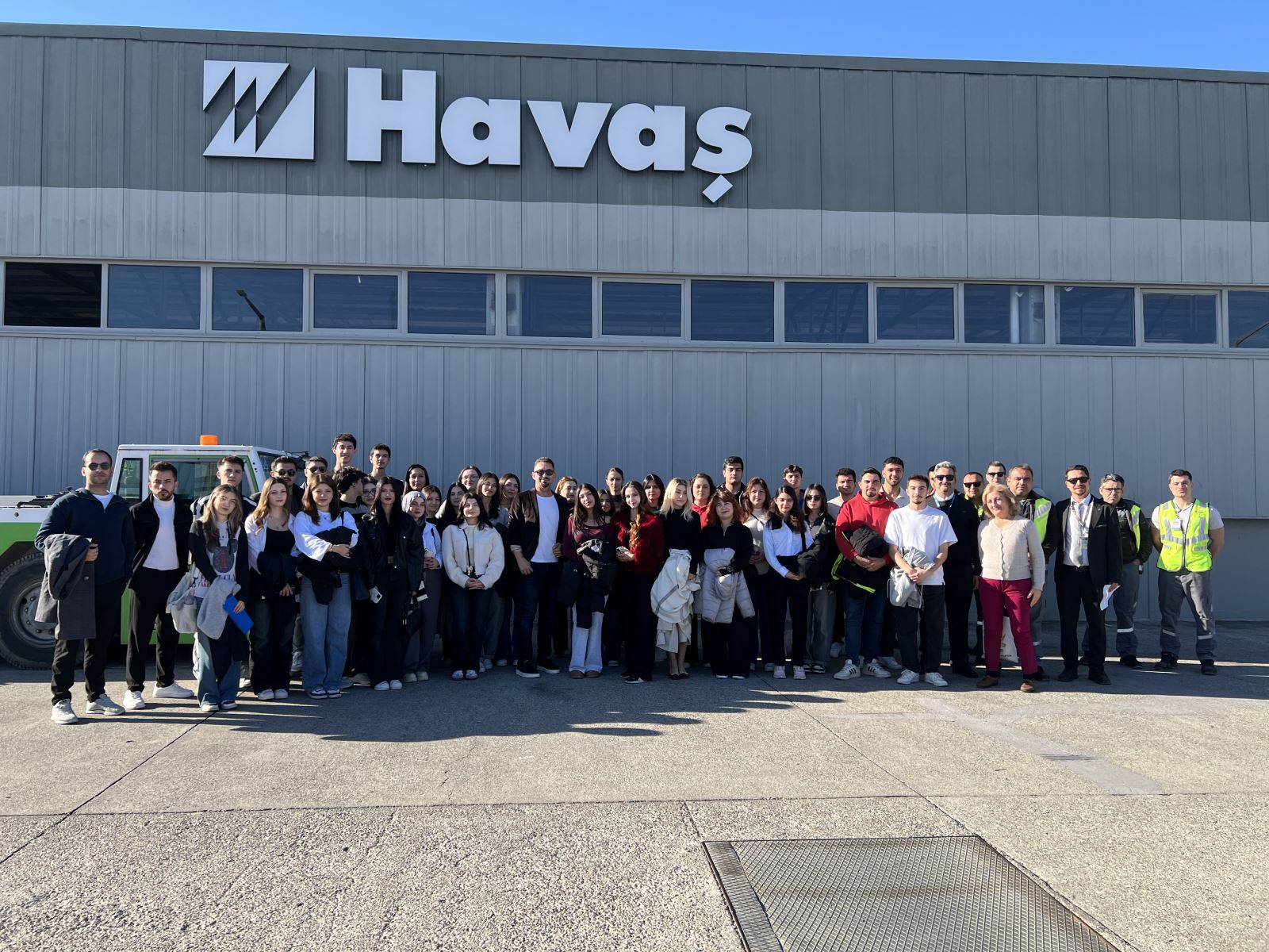 In this shot, the group is posing in front of a large, industrial grey hangar with a prominent "Havaş" sign. The building has a long row of horizontal windows. Part of an airport ground vehicle is visible on the left, and airport staff in high-visibility yellow vests are standing with the students on the right.